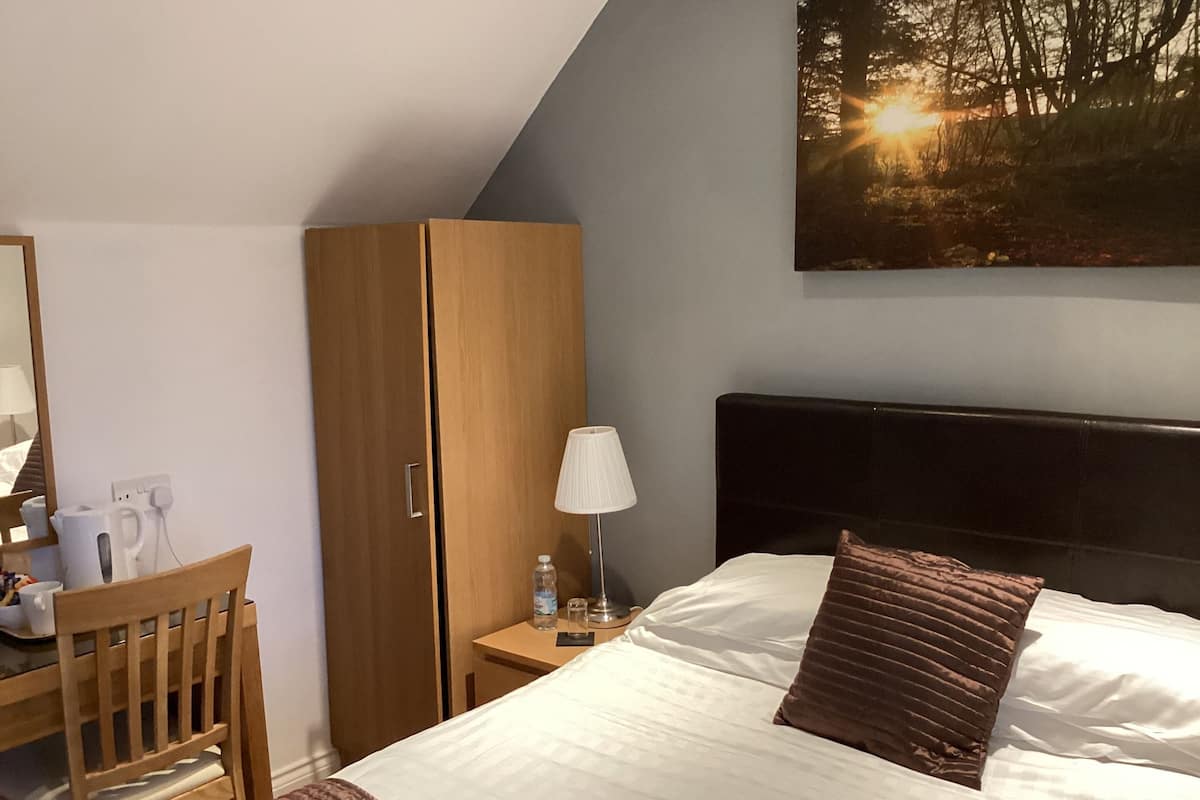 A hotel room with a bed, a wooden wardrobe, a desk with a chair, a lamp, and a painting of a forest scene.