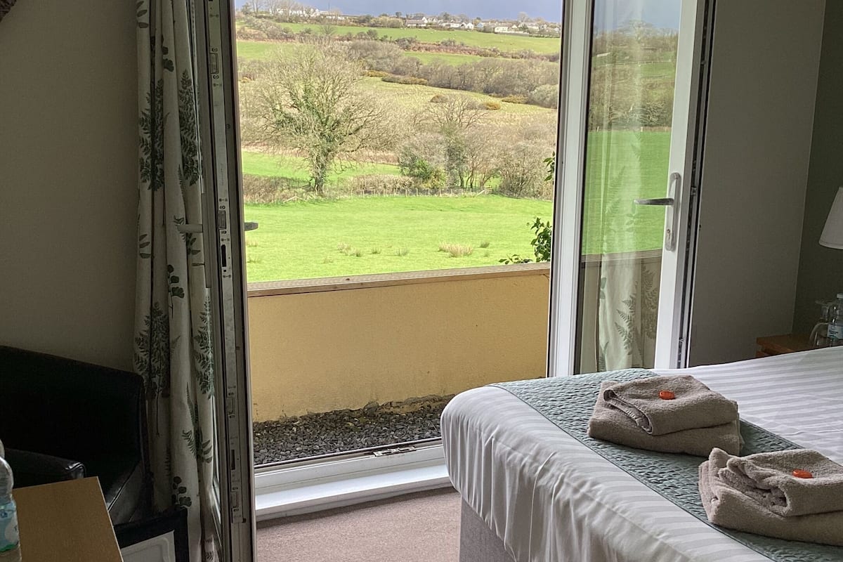 A bedroom with a bed, a desk, and a view of a green landscape through an open door.