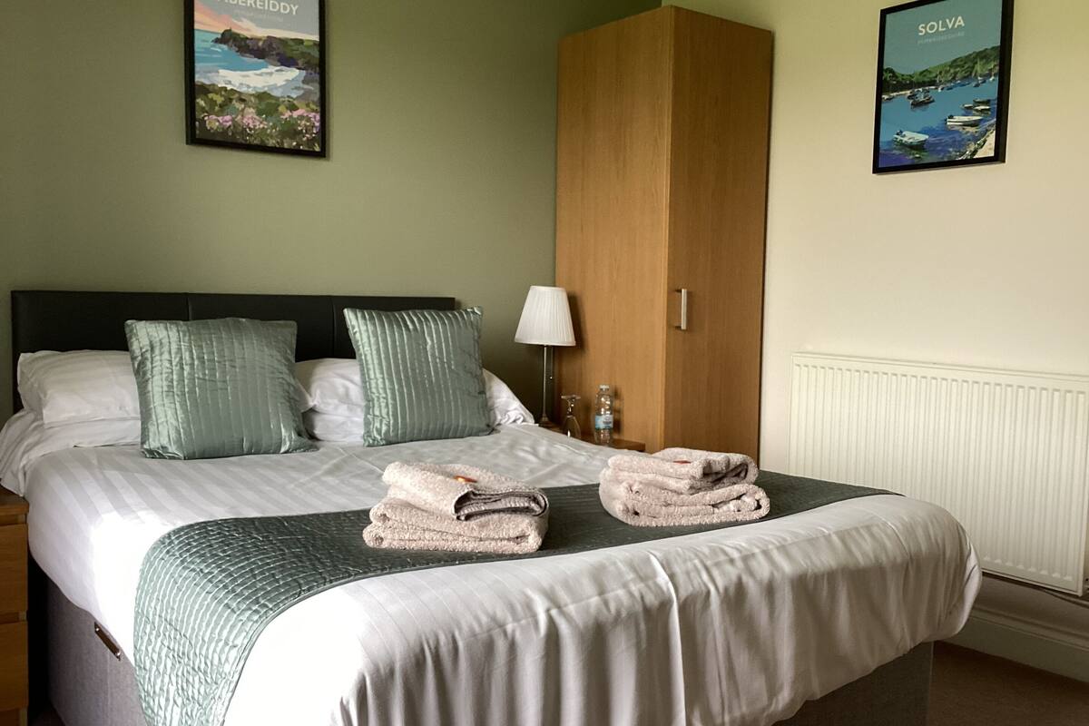 A neatly made bed with pillows and folded towels, a wooden wardrobe, and two framed pictures on the wall.