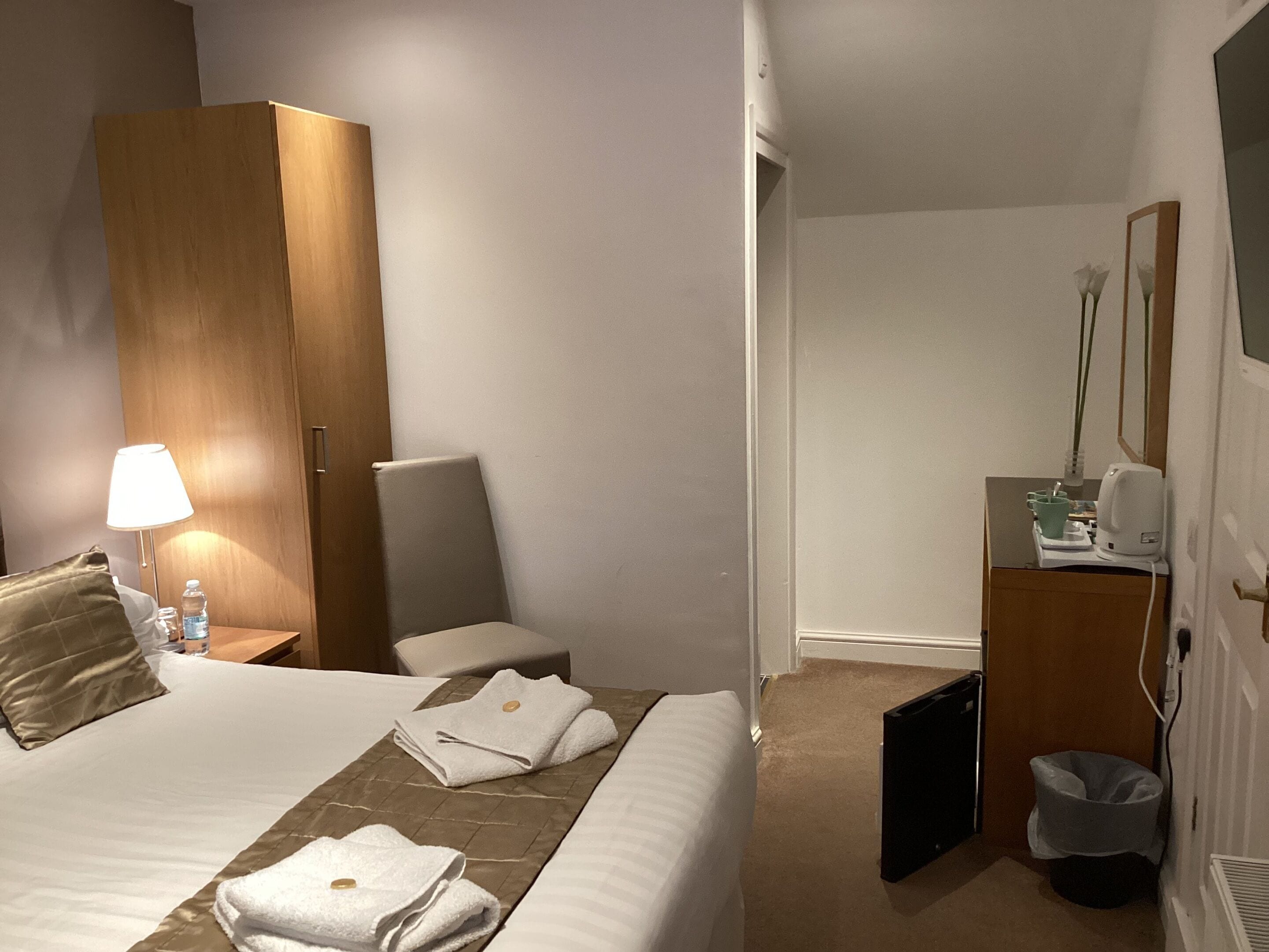 Et hotelværelse med en seng, et træskab, et skrivebord med lampe, en stol, en skraldespand og et lille bord med et spejl og en vase med blomster.