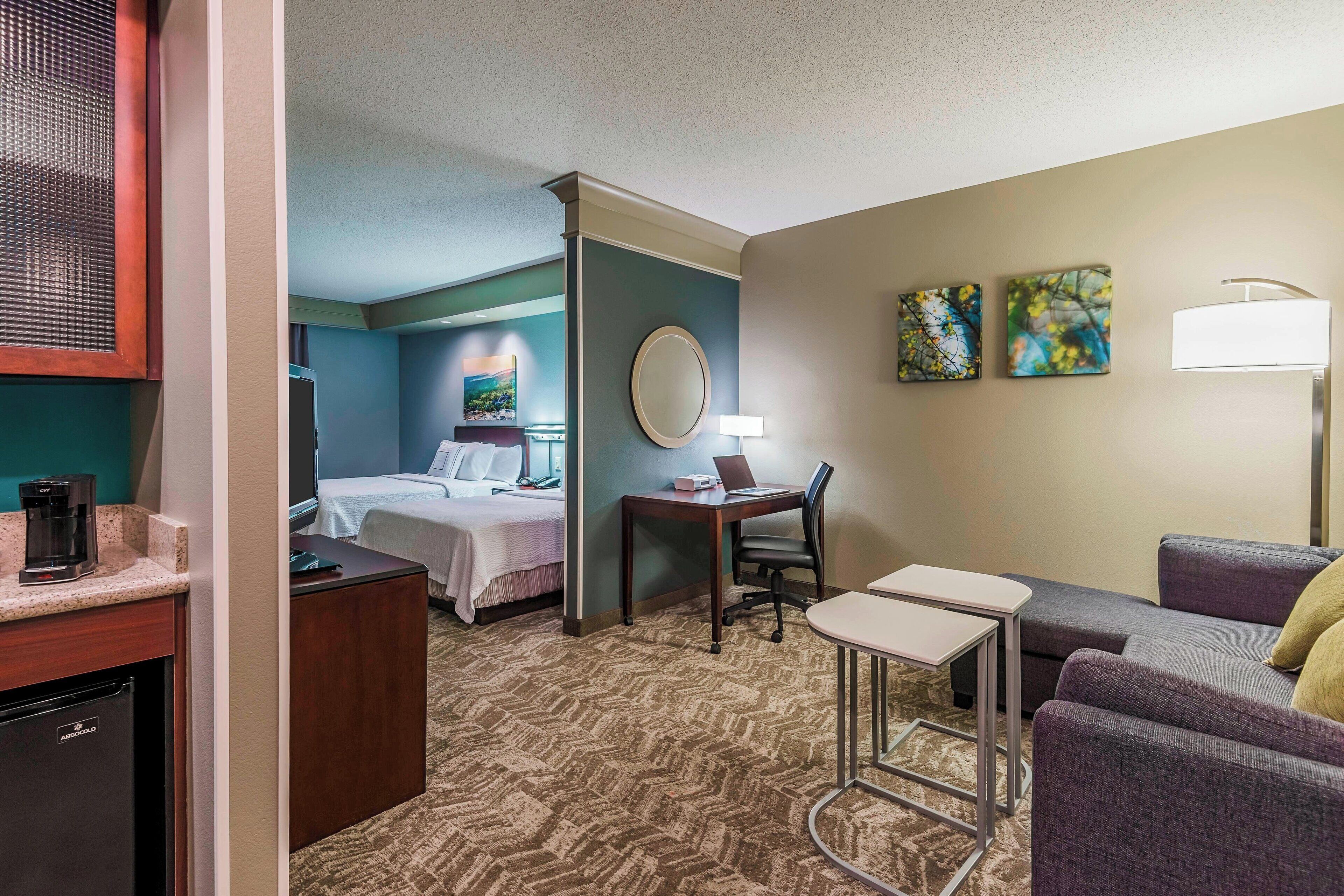 A hotel room with a bed, a desk with a chair, a small table, a sofa, a coffee machine, and a refrigerator.