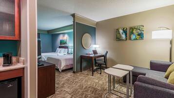 A hotel room with a bed, a desk with a chair, a small table, a sofa, a coffee machine, and a refrigerator.