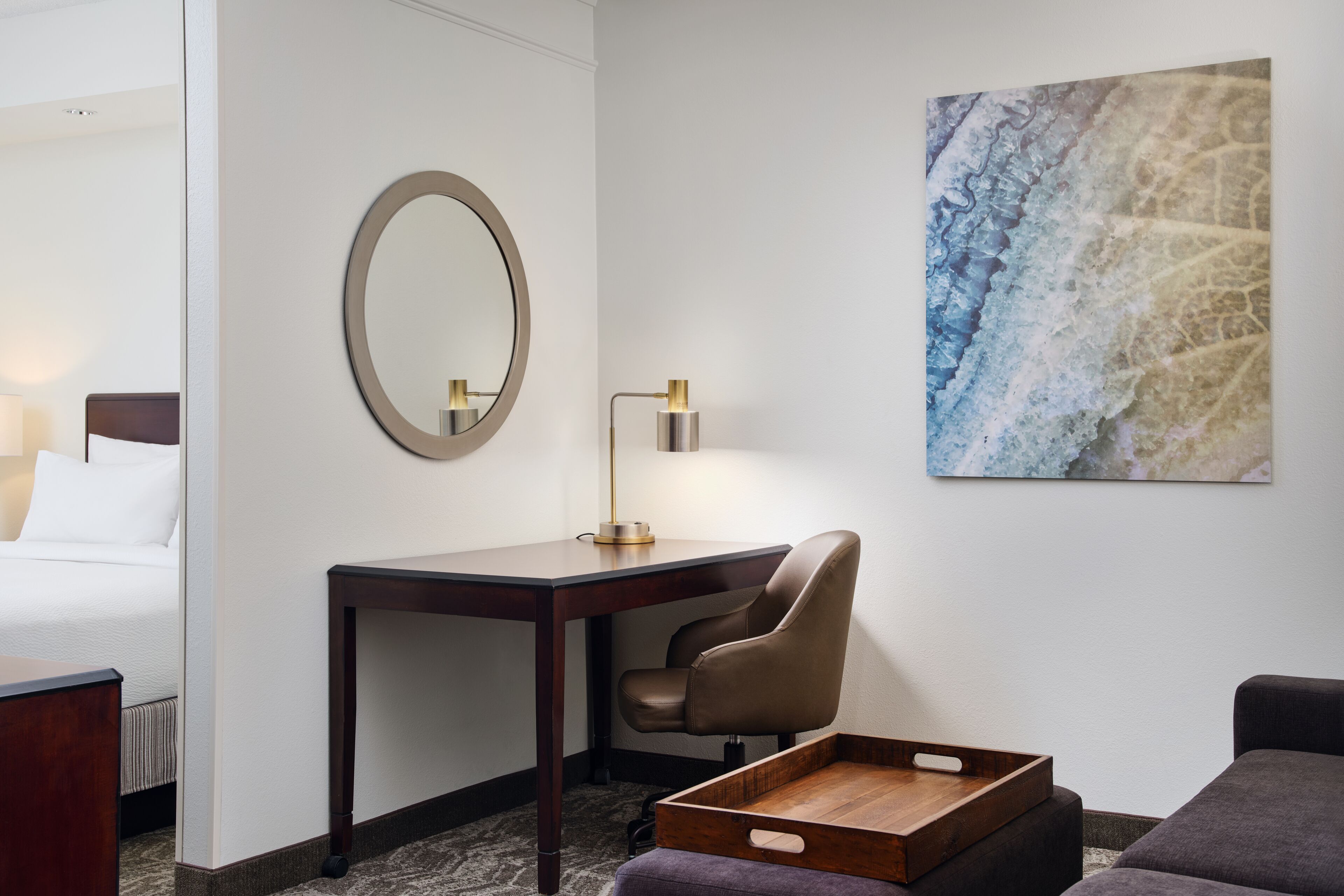 A hotel room with a bed, a desk with a chair, a desk lamp, a wooden side table, and a wall-mounted mirror.