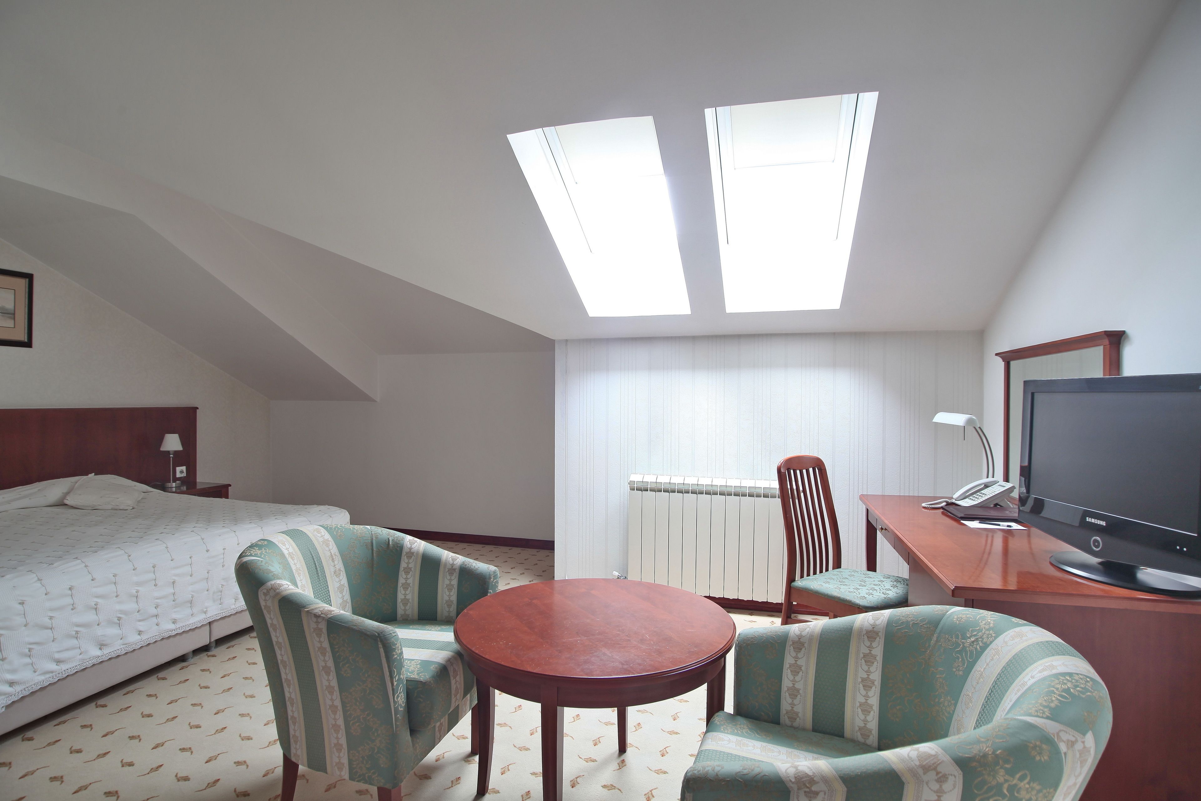 Habitación de hotel con una cama, dos sillones, una mesita, un escritorio con una computadora y un televisor. Hay tragaluces en el techo.