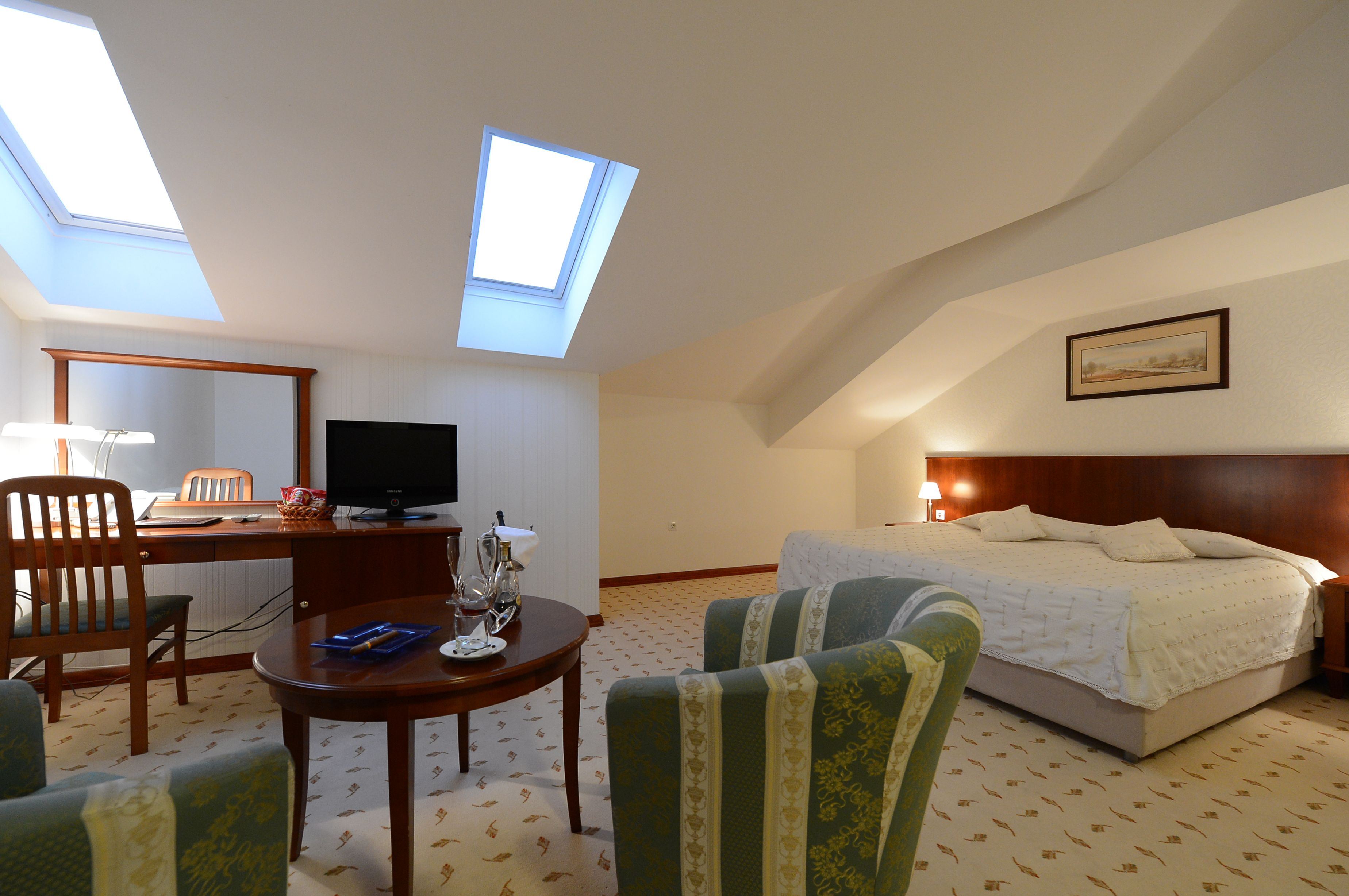 Una habitación de hotel con tragaluces, una cama, un escritorio con computadora, una silla y una mesita.