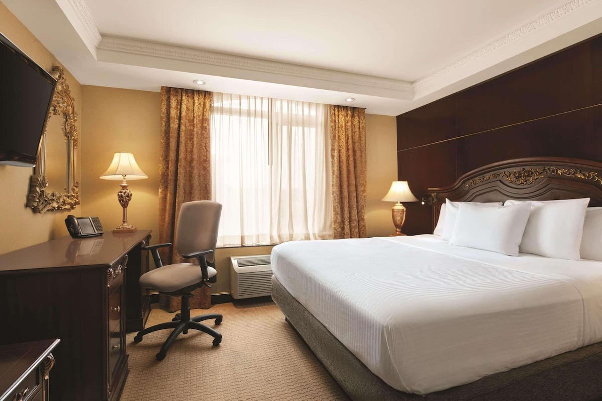 A hotel room with a large bed, a desk with a telephone, a chair, two lamps, and a television on the wall.