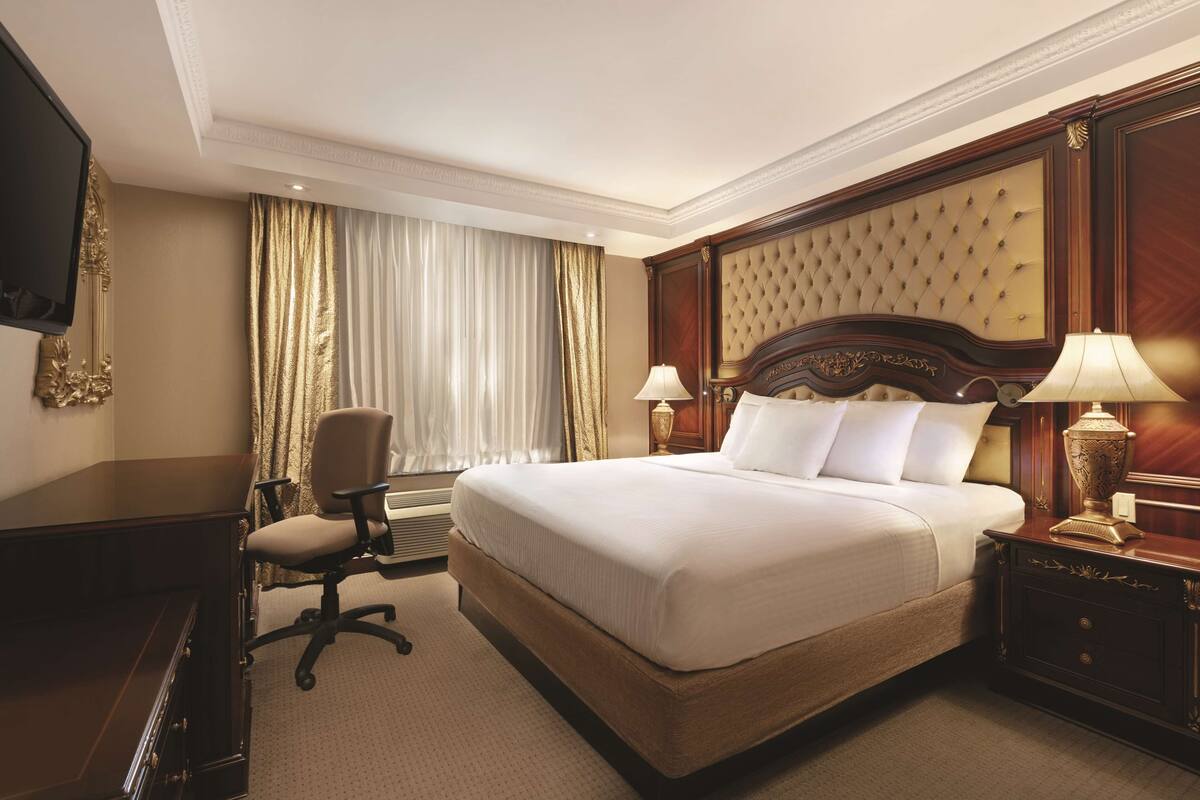 A hotel room with a large bed, a desk, a chair, a television, and a nightstand with a lamp.