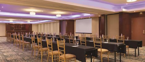 A conference room with rows of tables and chairs, a projector screen, and a carpeted floor.