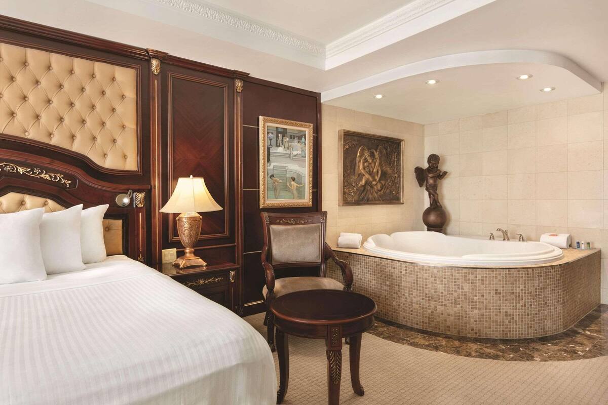 A hotel room with a large bed, a sitting area with a chair, a round bathtub, and a painting on the wall.