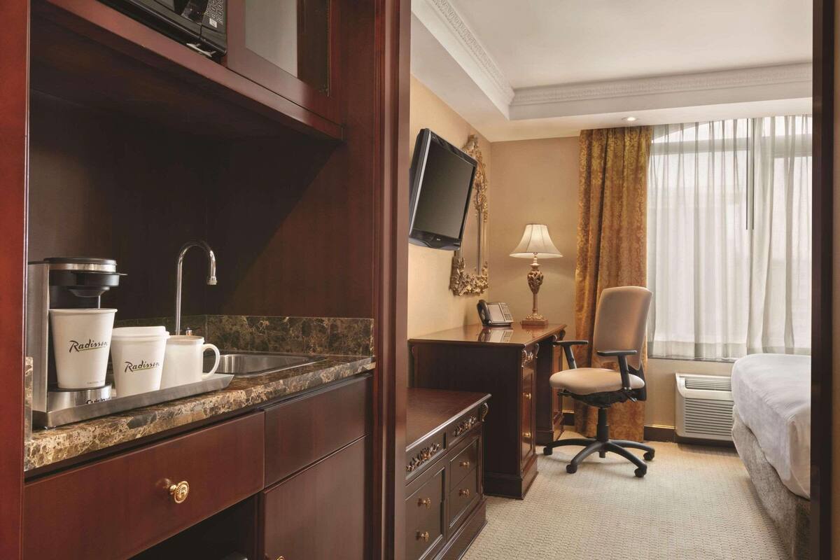A hotel room with a marble countertop, a sink, a desk with a chair, a television, and a bed.