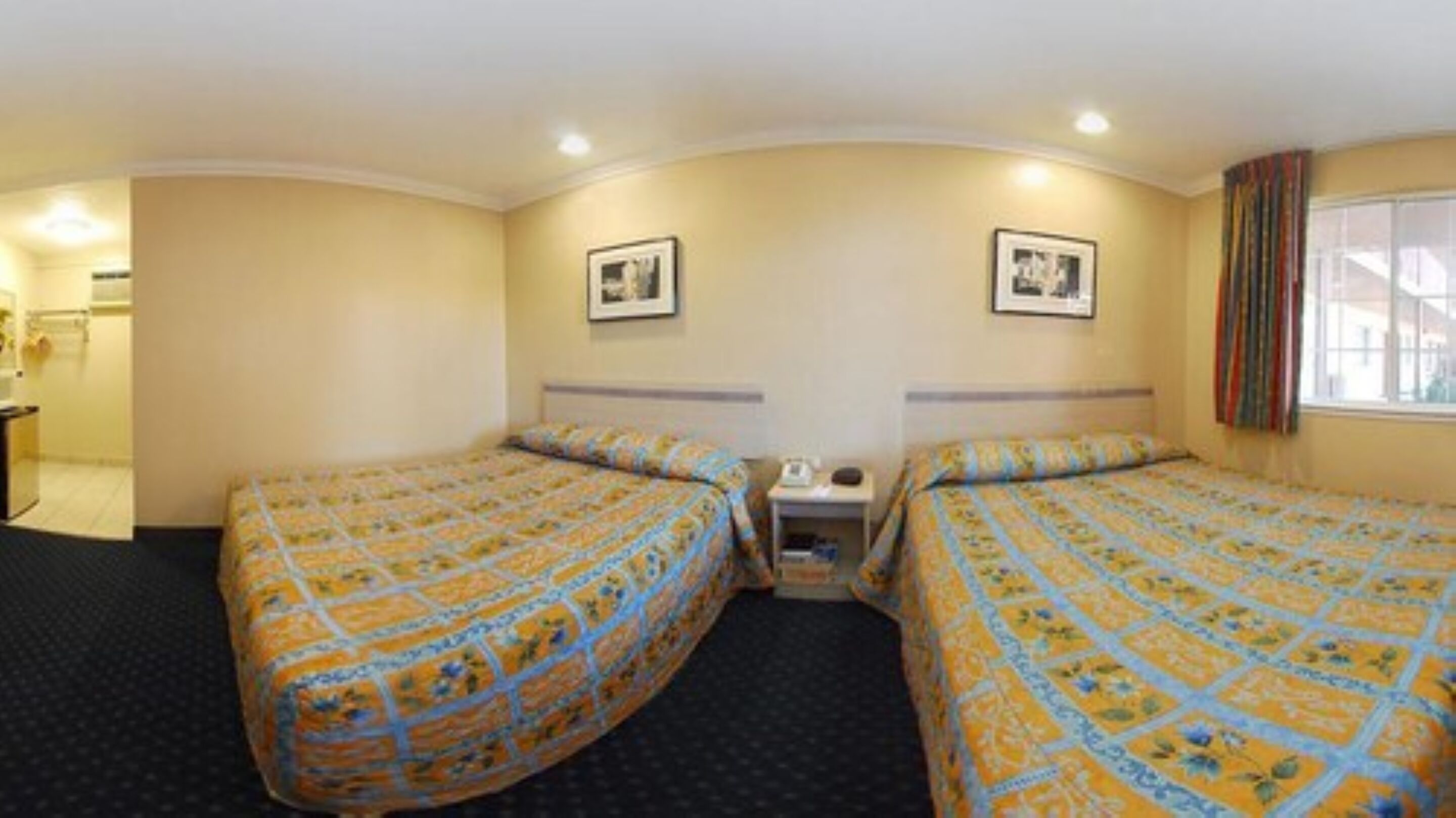 A hotel room with two beds, a small bedside table with a lamp, a window with curtains, and a picture on the wall.