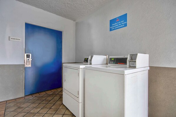 A laundry area with two washing machines, a blue door labeled "Boiler Room," and a sign stating "Studio 6 is not responsible for items left unattended."