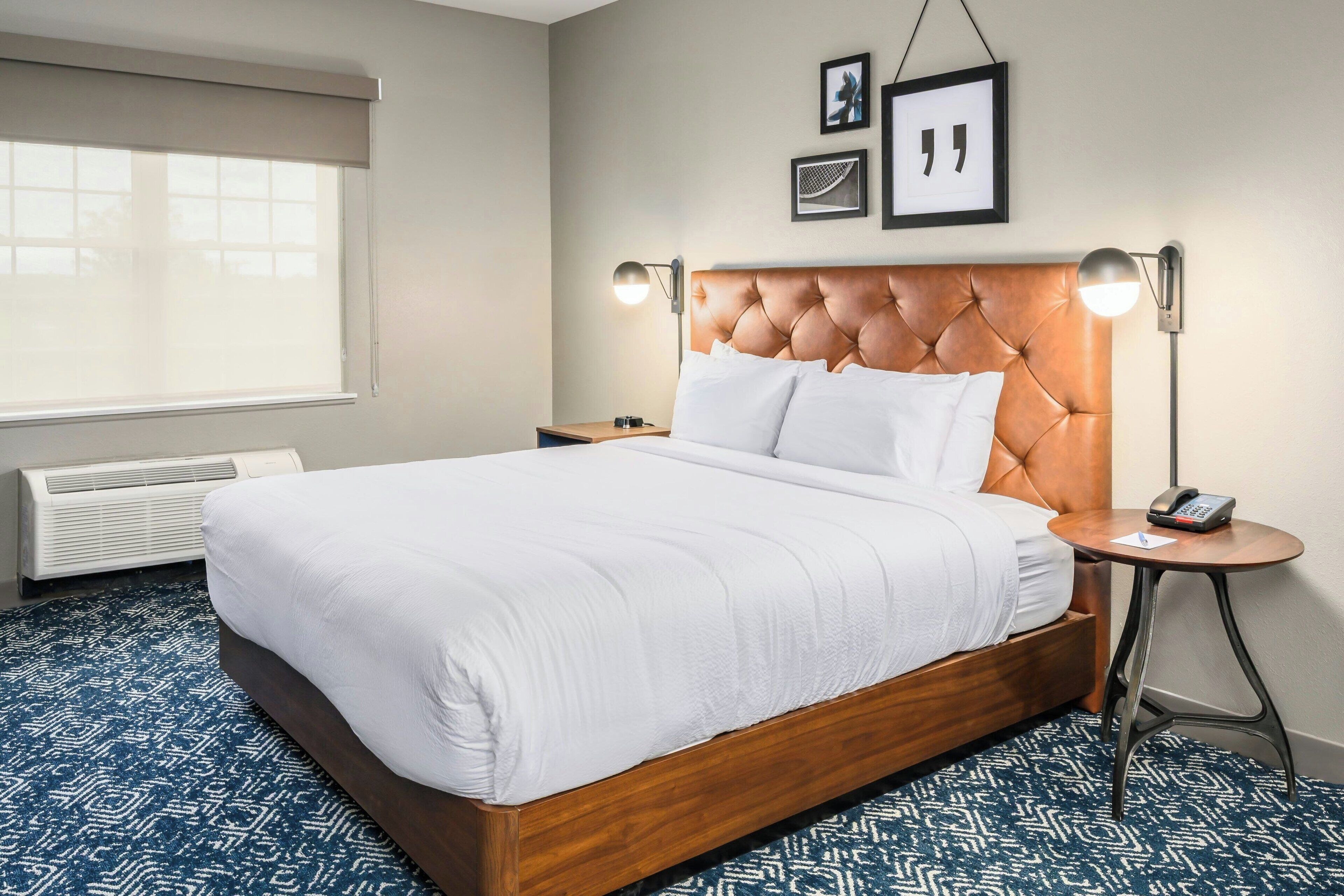 A hotel room with a large bed, a bedside table with a lamp, a phone, and a framed picture on the wall.