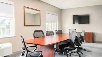 A conference room with a wooden table, six black office chairs, a large mirror, a flat-screen TV, and a window with blinds.