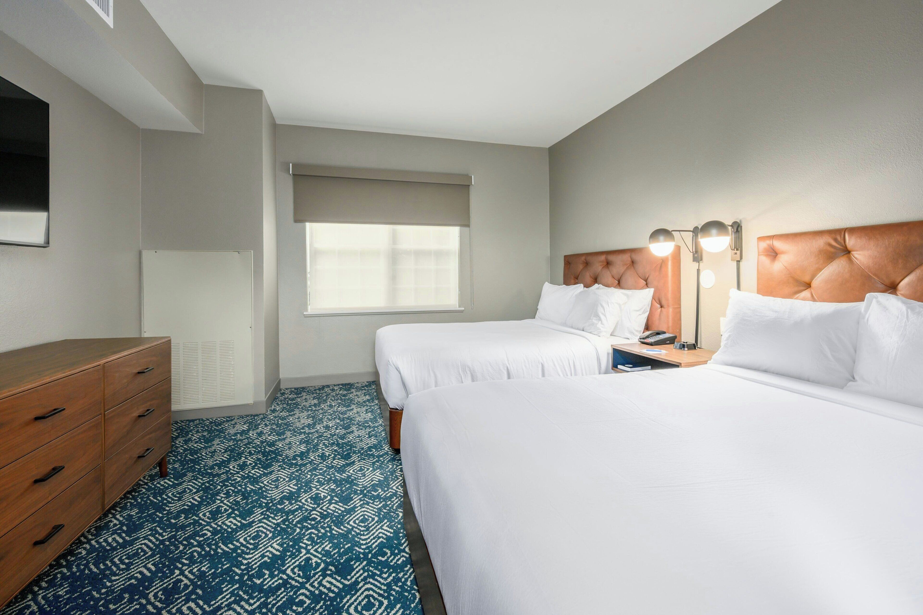 A hotel room with two beds, a wooden dresser, a window with blinds, and a wall-mounted TV.