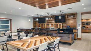 Un moderno vestíbulo de hotel con una mesa de comedor de madera en el centro, varias zonas de estar, un área de cocina y pantallas de televisión integradas.