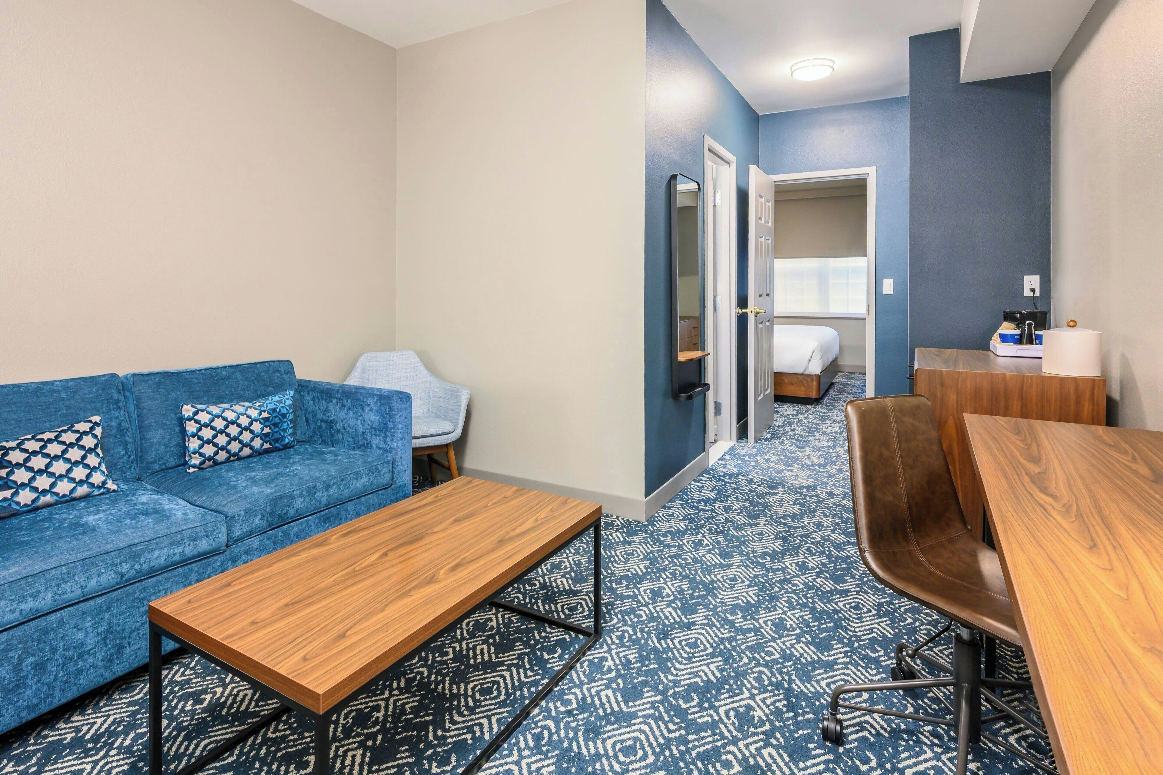 Ein modernes Hotelzimmer mit einem blauen Sofa, einem Holztisch, einem braunen Ledersessel und einem kleinen Schreibtisch mit Spiegel.