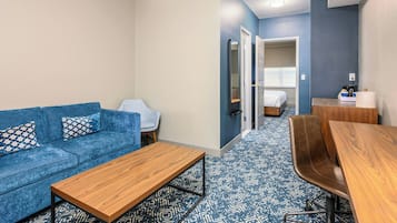 Ein modernes Hotelzimmer mit einem blauen Sofa, einem Holztisch, einem braunen Ledersessel und einem kleinen Schreibtisch mit Spiegel.