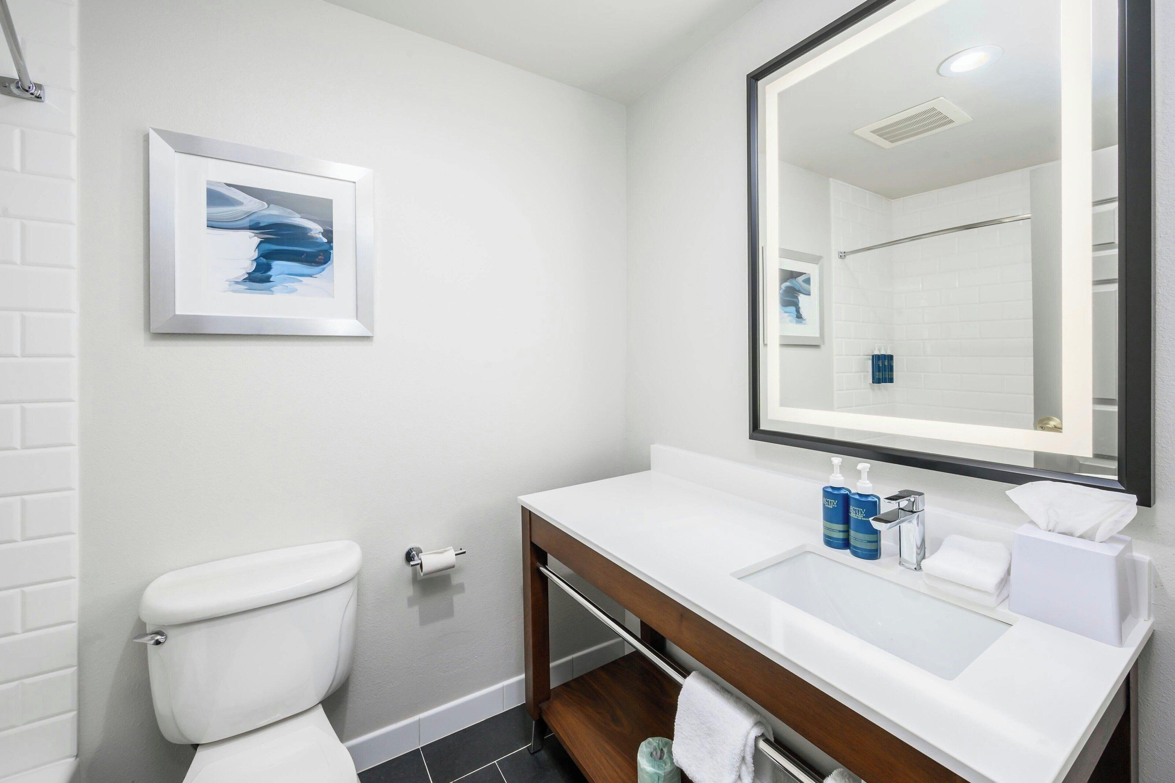 A modern bathroom with a white toilet, a framed abstract painting, a large mirror, and a white sink with toiletries.