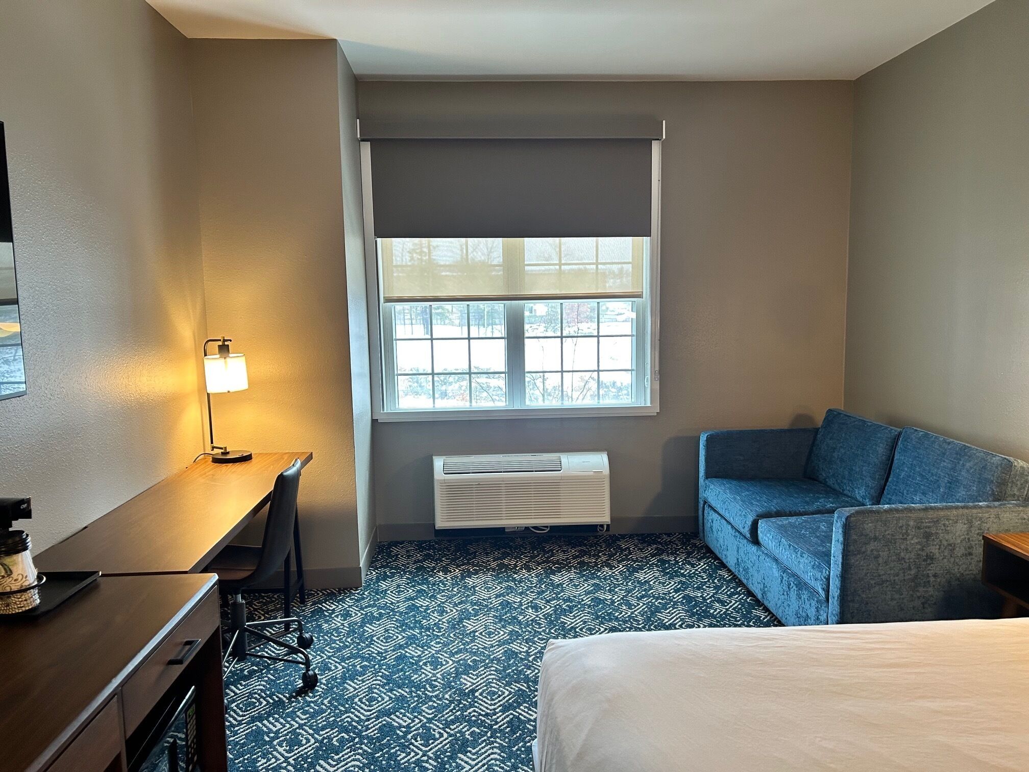 A hotel room with a desk, a bed, a blue couch, a window with blinds, and an air conditioning unit.