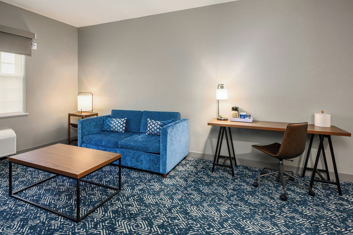 A room with a blue sofa, a wooden coffee table, a desk with a chair, and a patterned carpet.