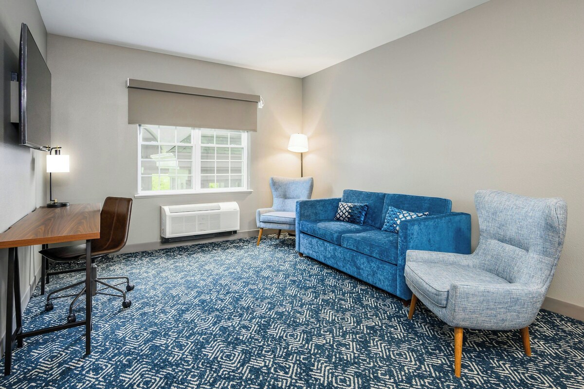 A room with a blue patterned carpet, a blue sofa, a grey armchair, a wooden desk, a chair, a lamp, and a window with blinds.