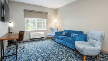 Una habitación con alfombra azul estampada, un sofá azul, un sillón gris, un escritorio de madera, una silla, una lámpara y una ventana con persianas.