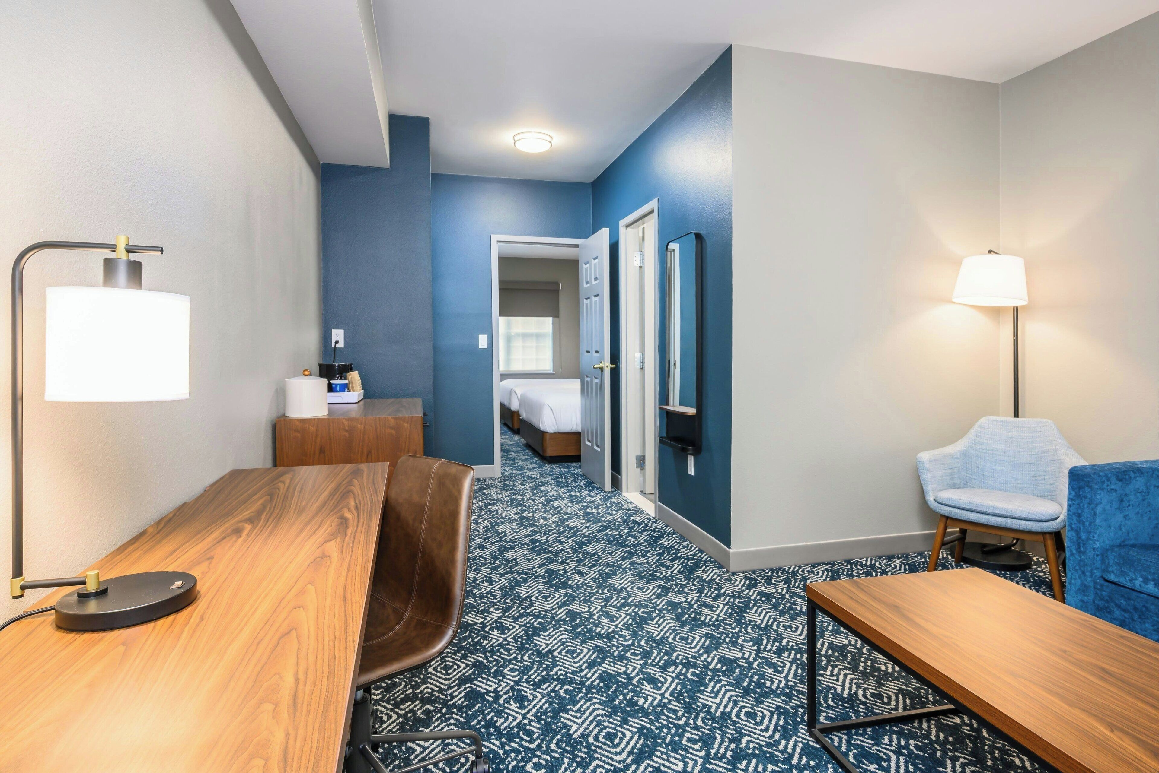 Een hotelkamer met een houten bureau, een blauw gestreept tapijt, een blauwe fauteuil, een nachtkastje en een bed met witte lakens.