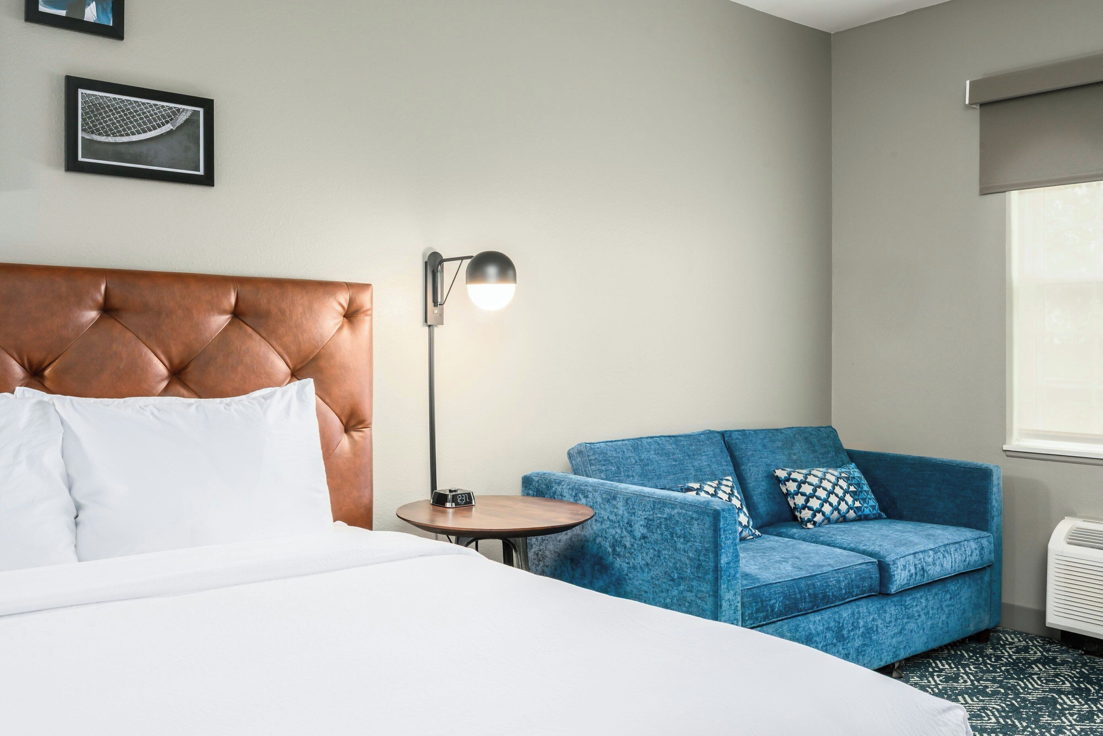 Quarto de hotel com cama e cabeceira estofada, um sofá azul com almofadas estampadas, uma mesinha redonda com abajur e um quadro na parede.