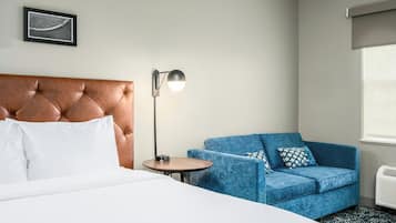 Quarto de hotel com cama e cabeceira estofada, um sofá azul com almofadas estampadas, uma mesinha redonda com abajur e um quadro na parede.