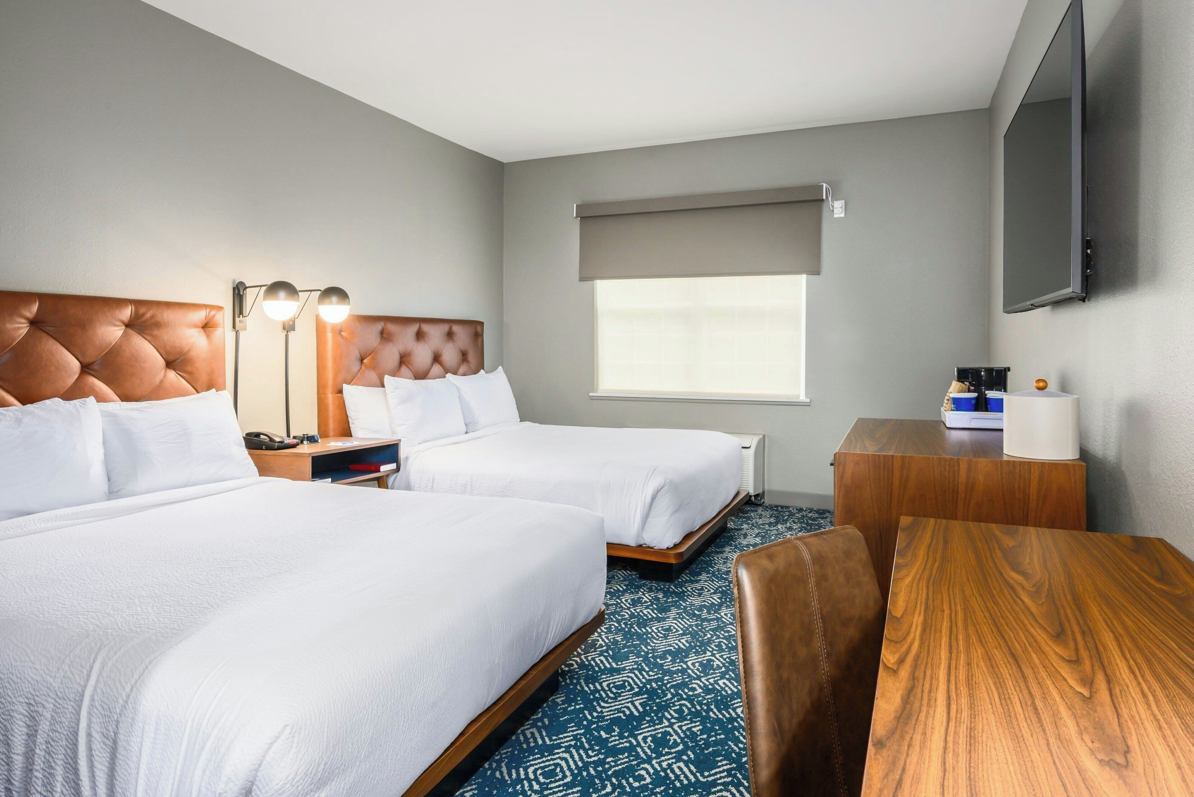 Quarto de hotel com duas camas, uma escrivaninha, uma cadeira, uma televisão e uma janela com vista.