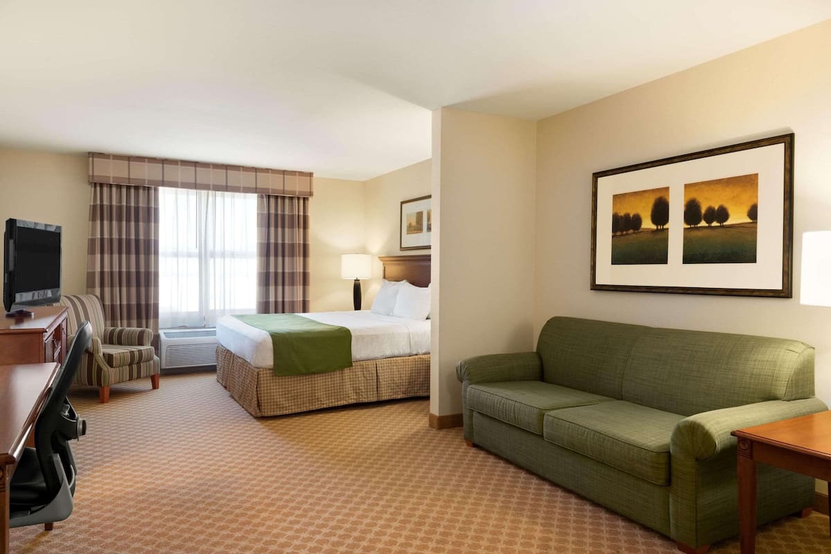 A hotel room with a bed, a green sofa, a desk with a chair, a TV, a lamp, and a painting on the wall.