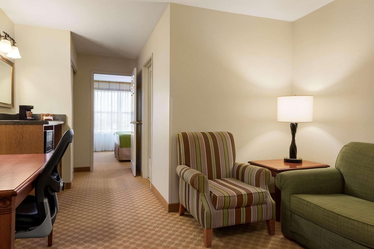 A hotel room with a dining table, a striped armchair, a green sofa, a lamp, and a view of a hallway with a door.