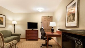 A hotel room with a green sofa, a wooden desk, a television, a microwave, a chair, a dresser, and a framed picture on the wall. - Country Inn & Suites by Radisson, Peoria North, IL (Peoria)