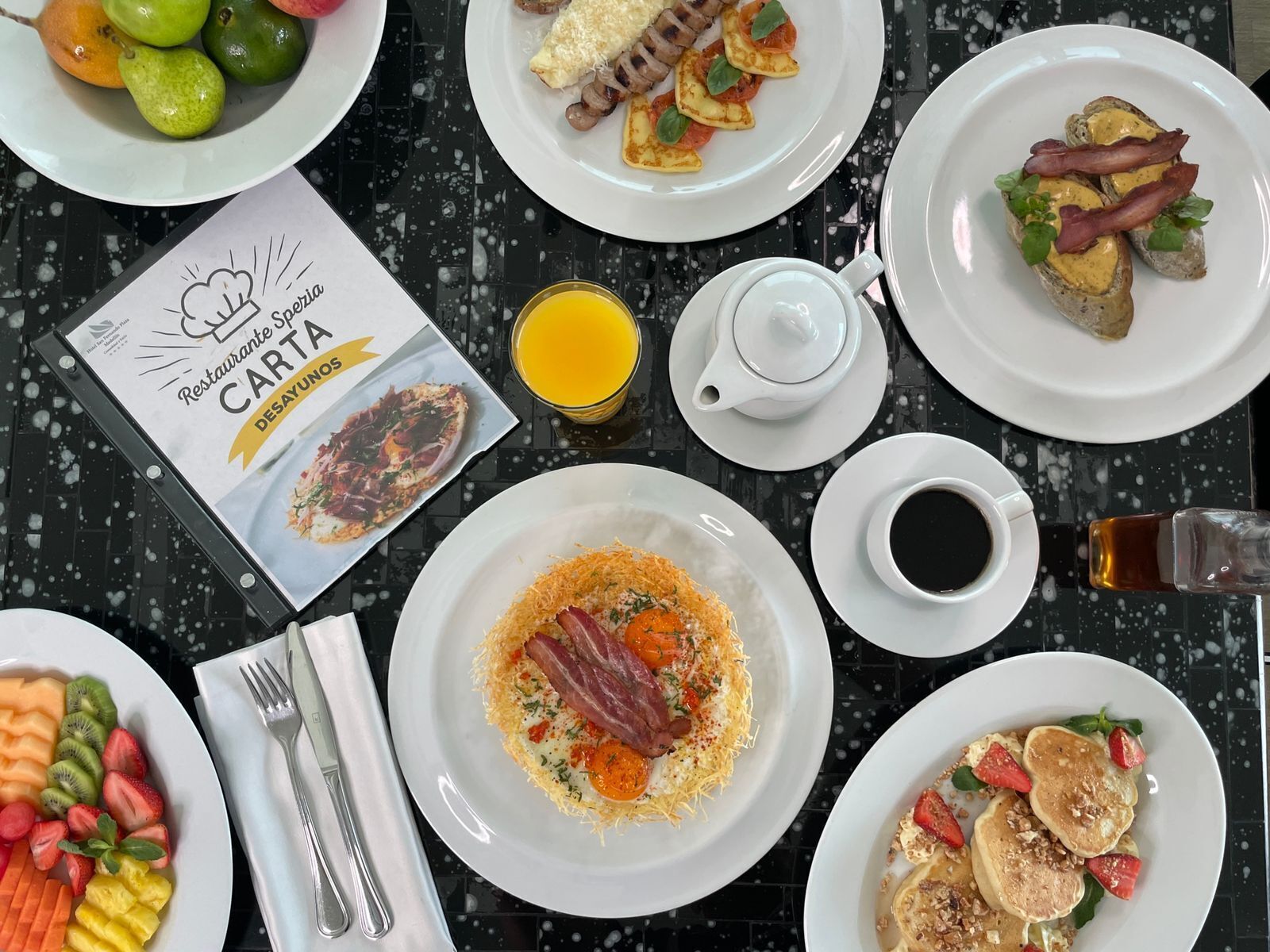Una mesa de desayuno con diversos platos, incluyendo huevos, tocino, panqueques y fruta. Se ve un menú que dice "Restaurante Sp. Carta Desayunos" y un vaso de jugo de naranja.
