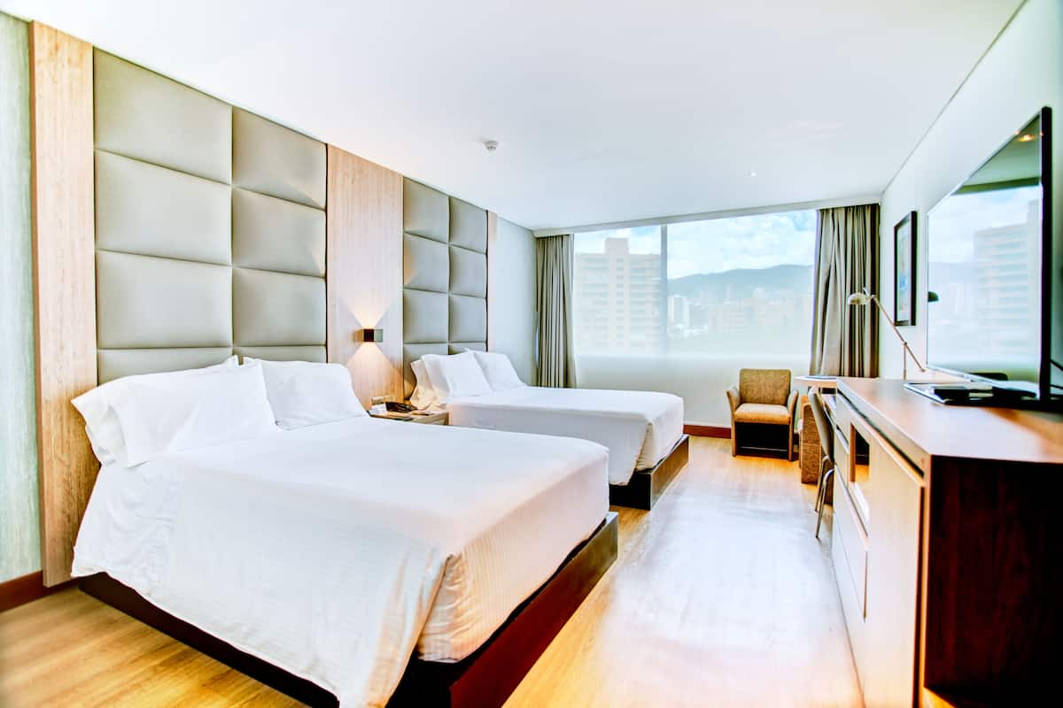 Ein Hotelzimmer mit zwei Betten, einem Schreibtisch, einem Sessel und einem großen Fenster mit Blick auf die Stadt.