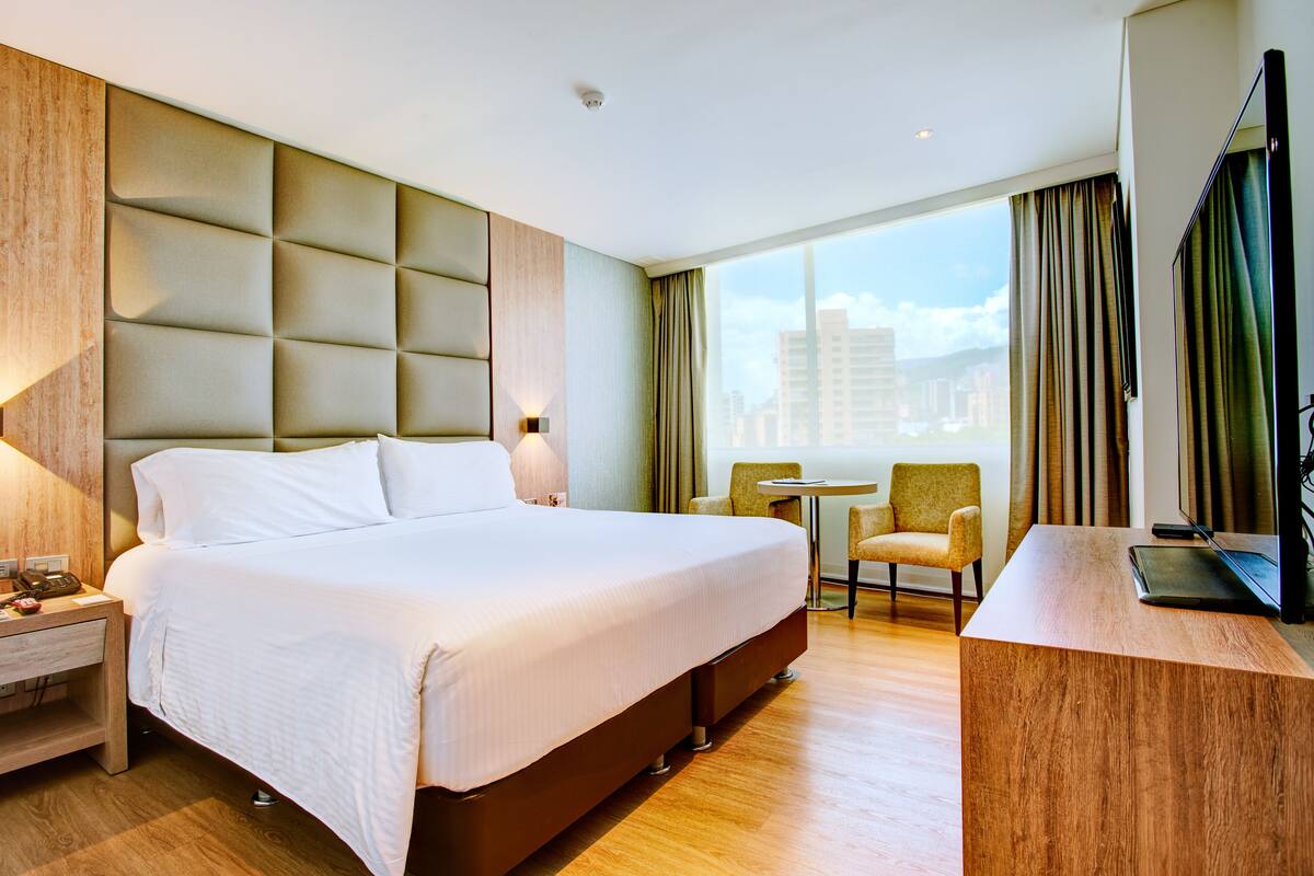 Ein Hotelzimmer mit einem großen Bett, einem Schreibtisch, einem Stuhl und Blick auf die Stadt durch das Fenster.