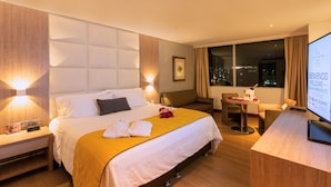 Habitación de hotel con una cama grande, lámparas de noche, un escritorio con computadora y vistas a la ciudad de noche.