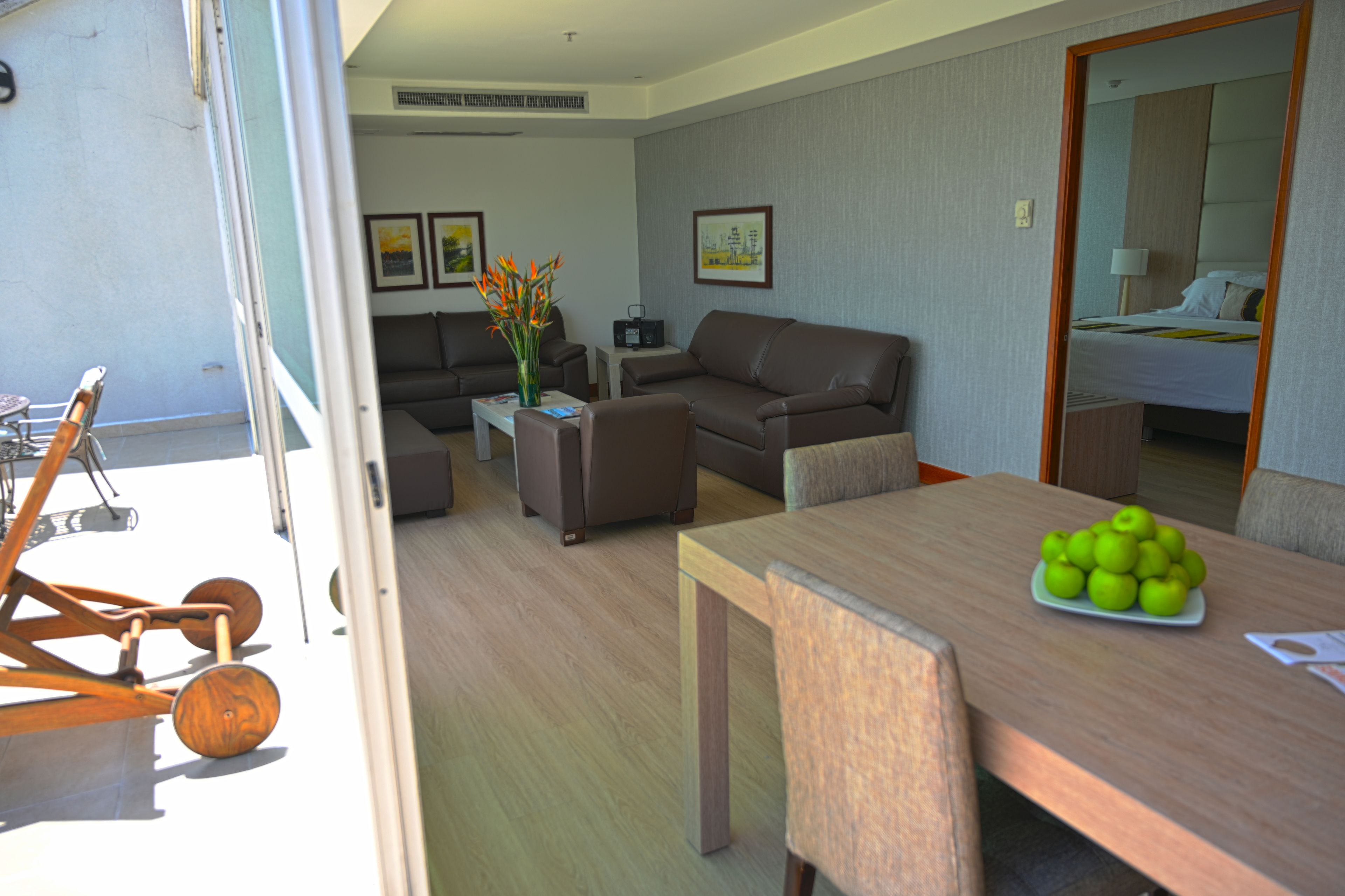 A modern hotel room with a sofa, a dining table with chairs, a small coffee table, and a decorative vase with flowers.