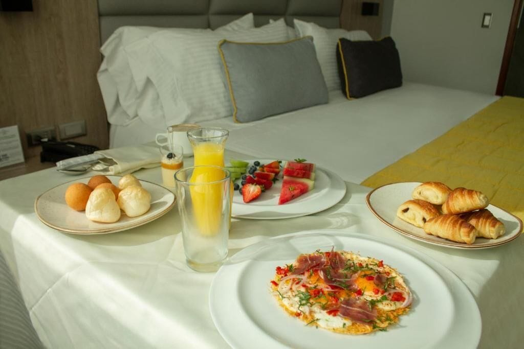 Un lit bien fait, avec des oreillers, une table de chevet sur laquelle se trouve un verre de jus d’orange, des assiettes de croissants, des fruits et un plat pour le petit-déjeuner.