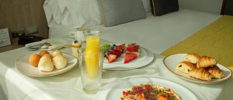 Un lit bien fait, avec des oreillers, une table de chevet sur laquelle se trouve un verre de jus d’orange, des assiettes de croissants, des fruits et un plat pour le petit-déjeuner.