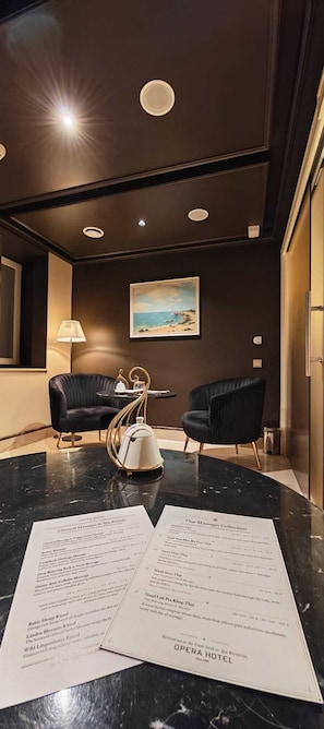 A hotel lobby with a marble reception desk, two black armchairs, a painting of a beach, and a menu on the desk. - Opera Hotel (Riga)
