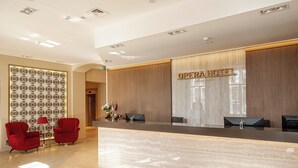Un hall d’accueil d’hôtel avec un comptoir en marbre, des chaises rouges, un mur décoratif et une enseigne indiquant « Hôtel Opéra ».