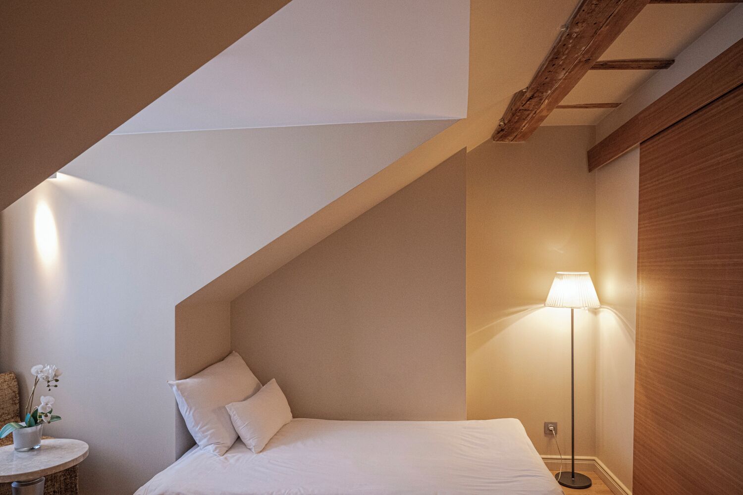 Een slaapkamer met een schuine kap, een bed met kussens, een nachtkastje met een lamp en een kleine plant.