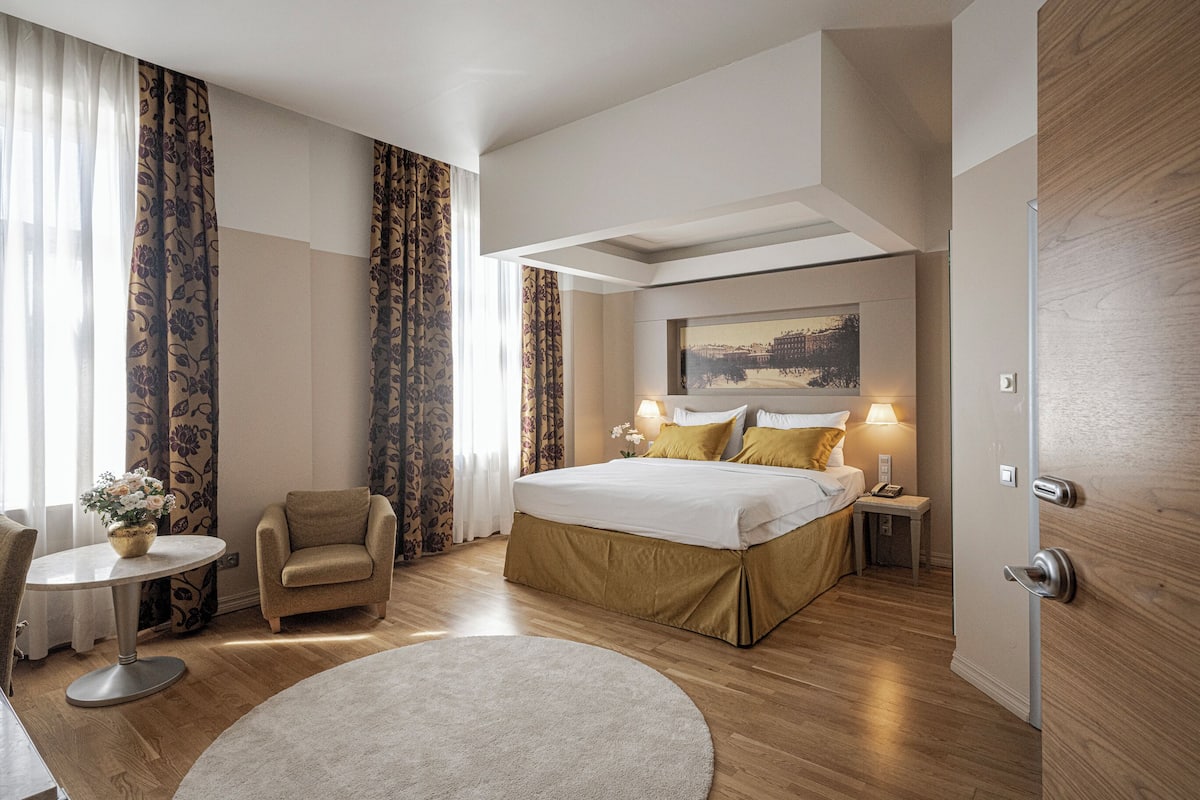 A hotel room with a bed, bedside table, lamp, chair, round table, and a painting on the wall.