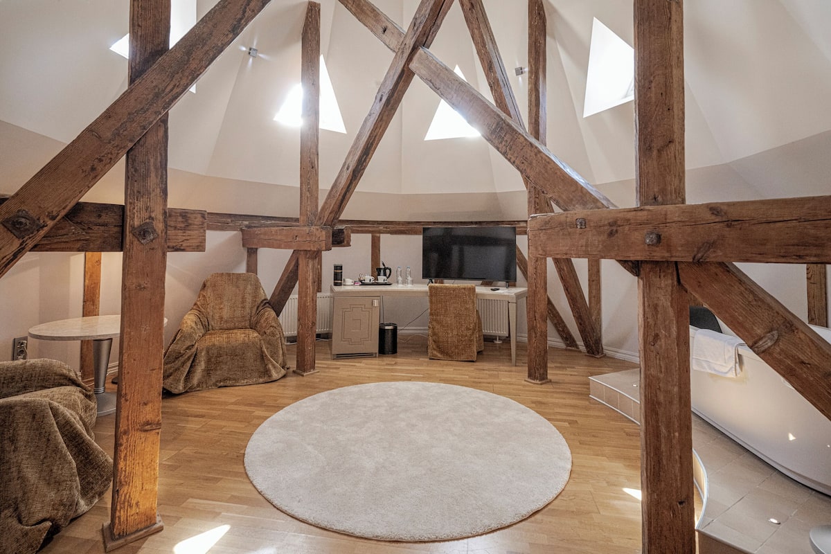 A spacious loft with exposed wooden beams, a round rug, seating areas, a kitchen, and a large TV.
