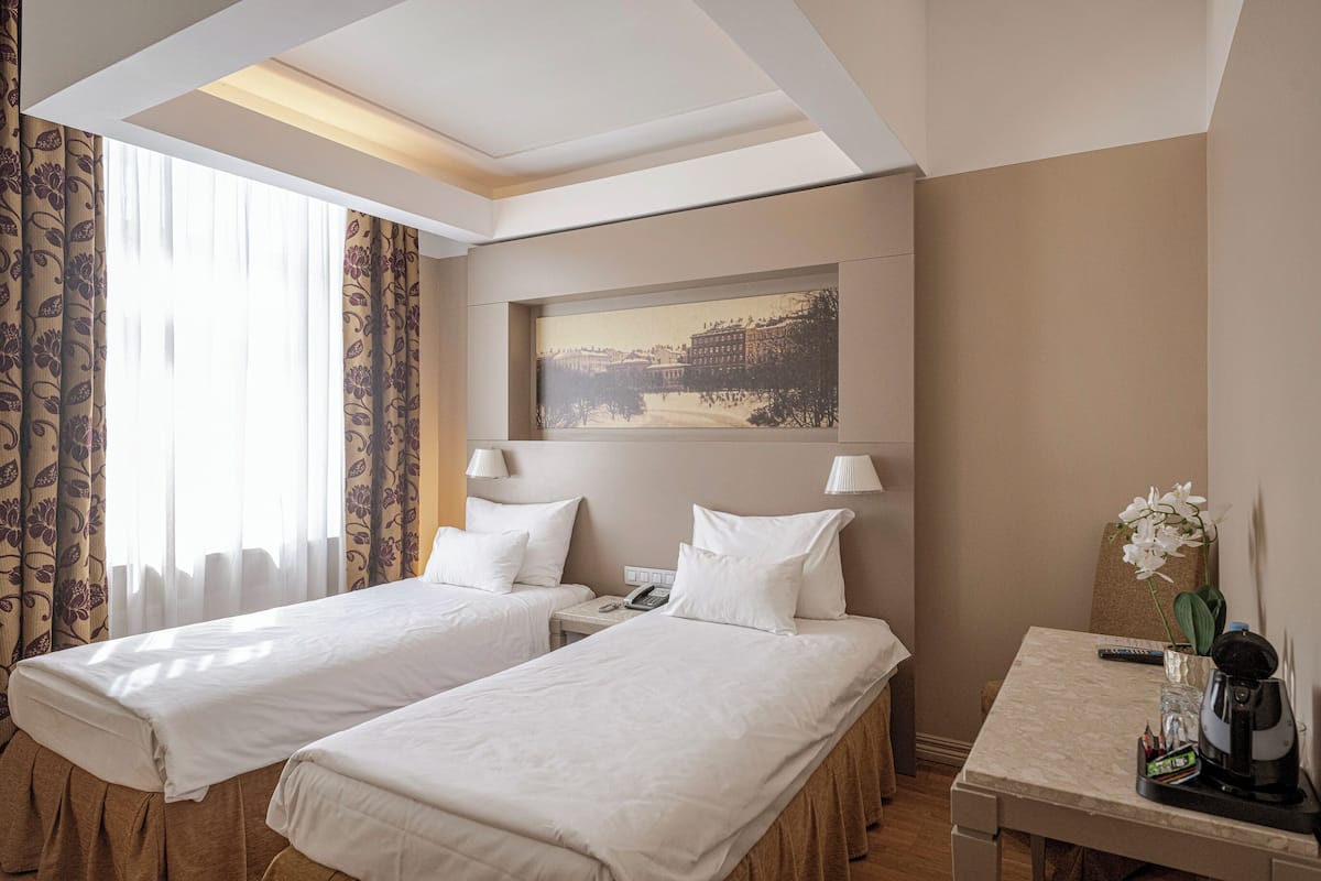 A hotel room with two beds, a desk, a telephone, a painting on the wall, and a window with curtains.