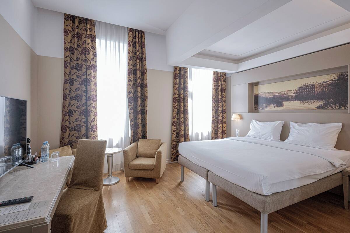 A hotel room with a bed, a desk with a chair, a TV, and a window with curtains.