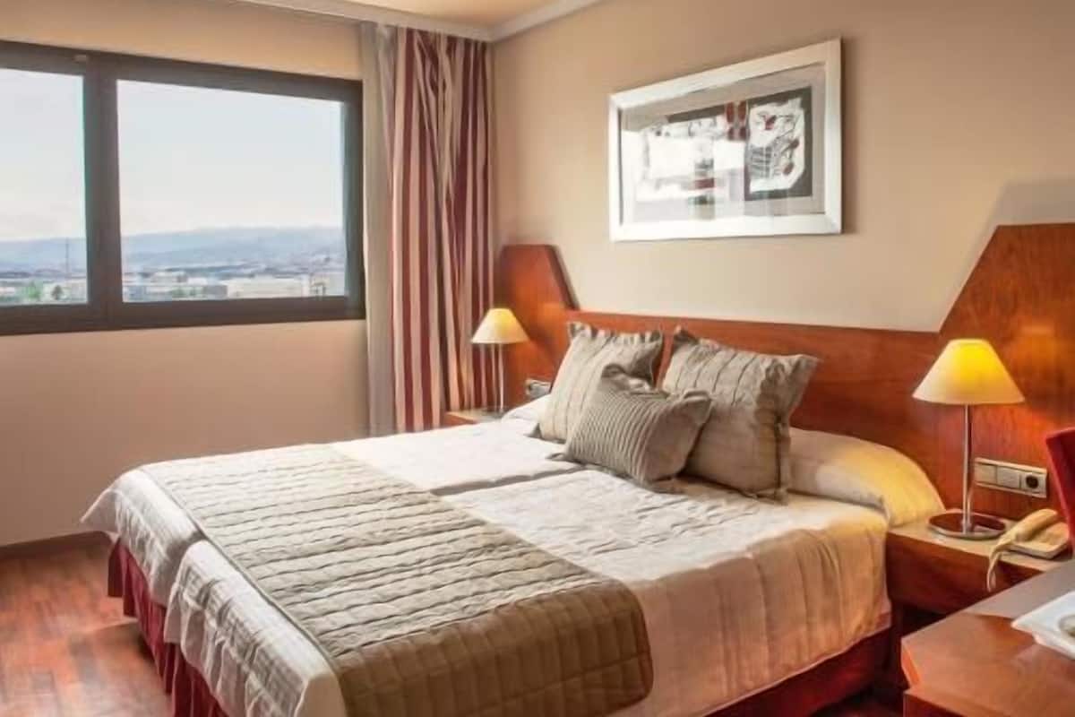 Ein Hotelzimmer mit einem Bett, zwei Nachttischlampen, einem fensterartigen Ausblick mit Vorhängen, einem gerahmten Bild an der Wand und einem Holzkopfende.