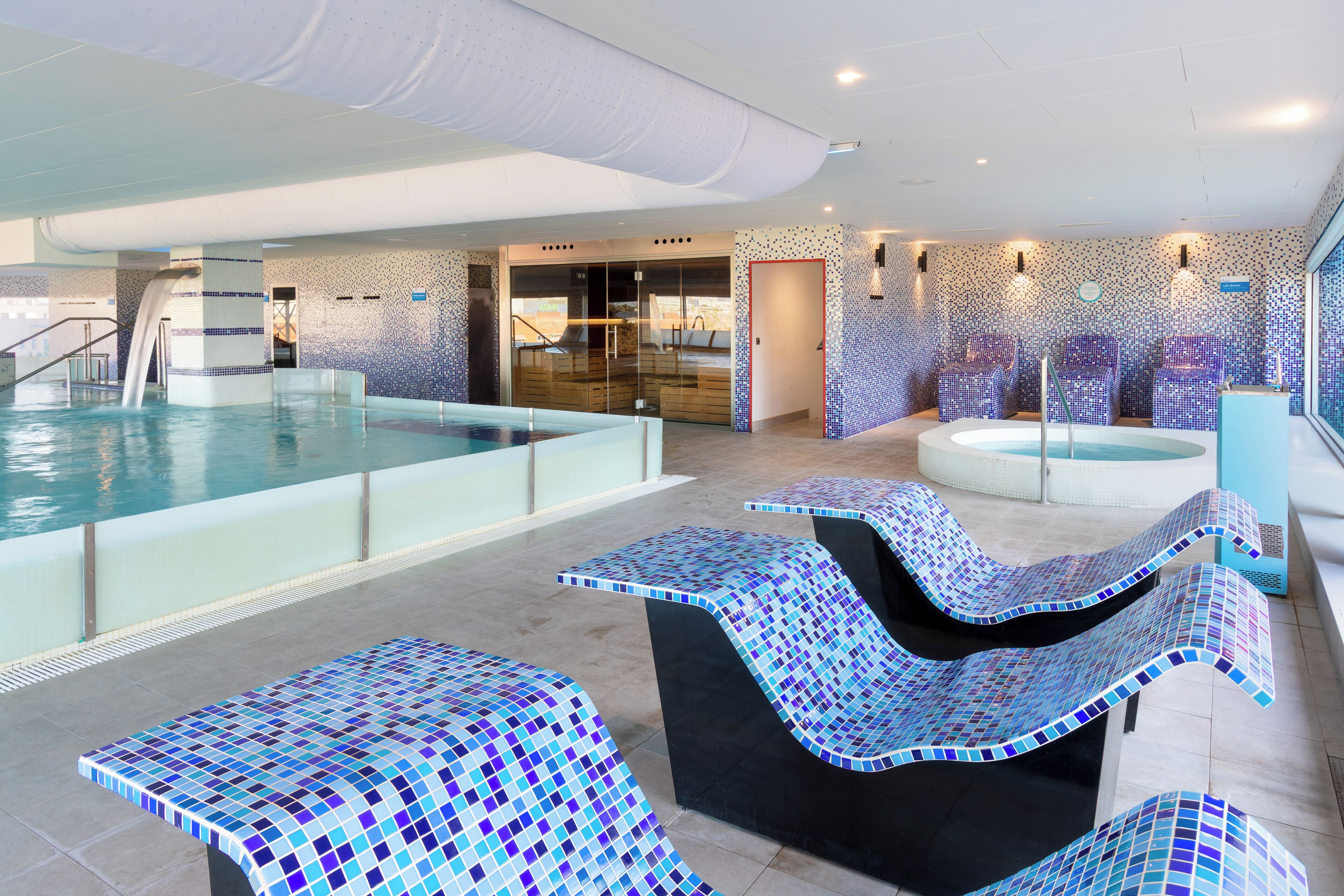 Area piscina interna con parete di vetro, vasca idromassaggio e sedute in mosaico.